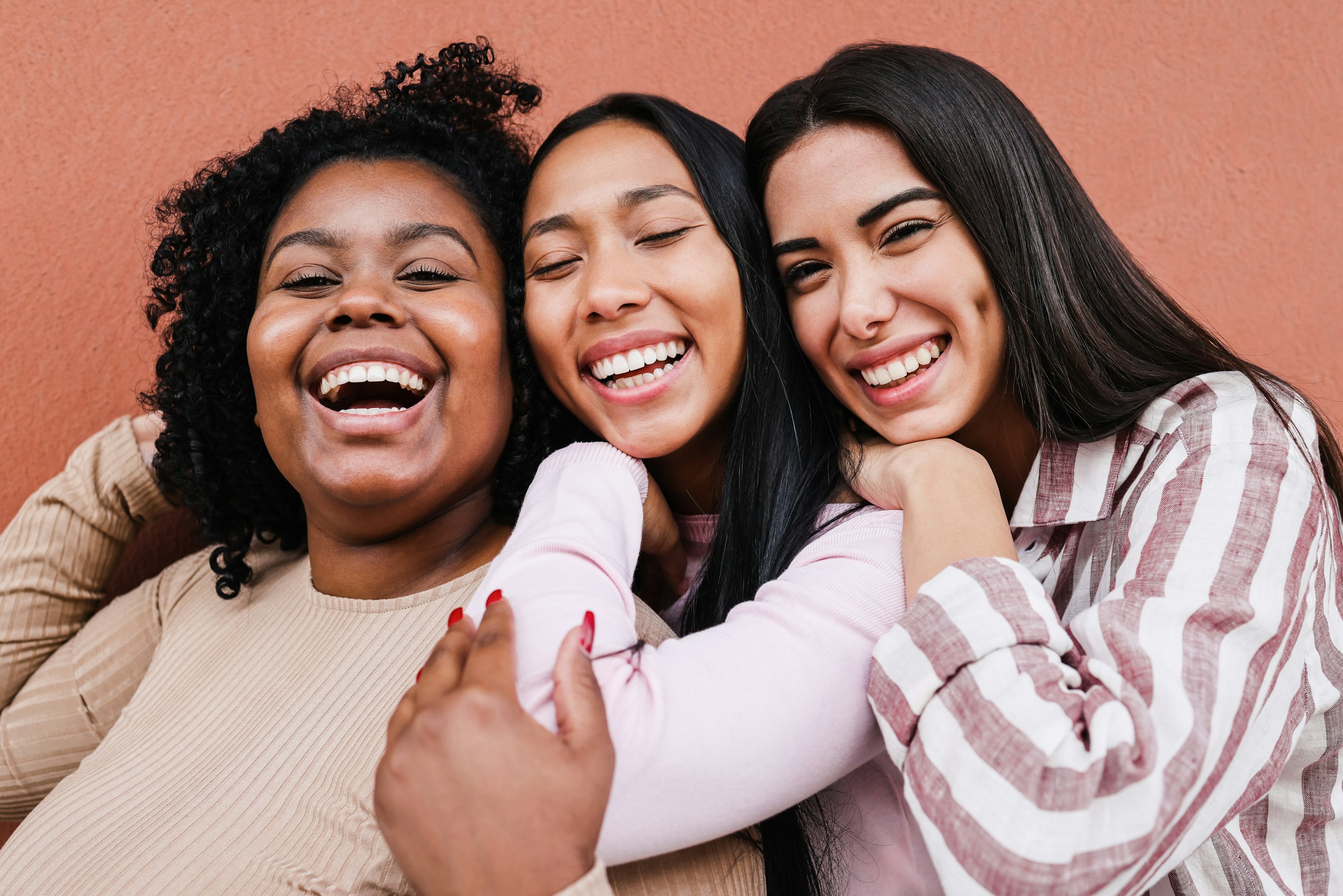Three women friends hugging and laughing against a warm terracotta wall
